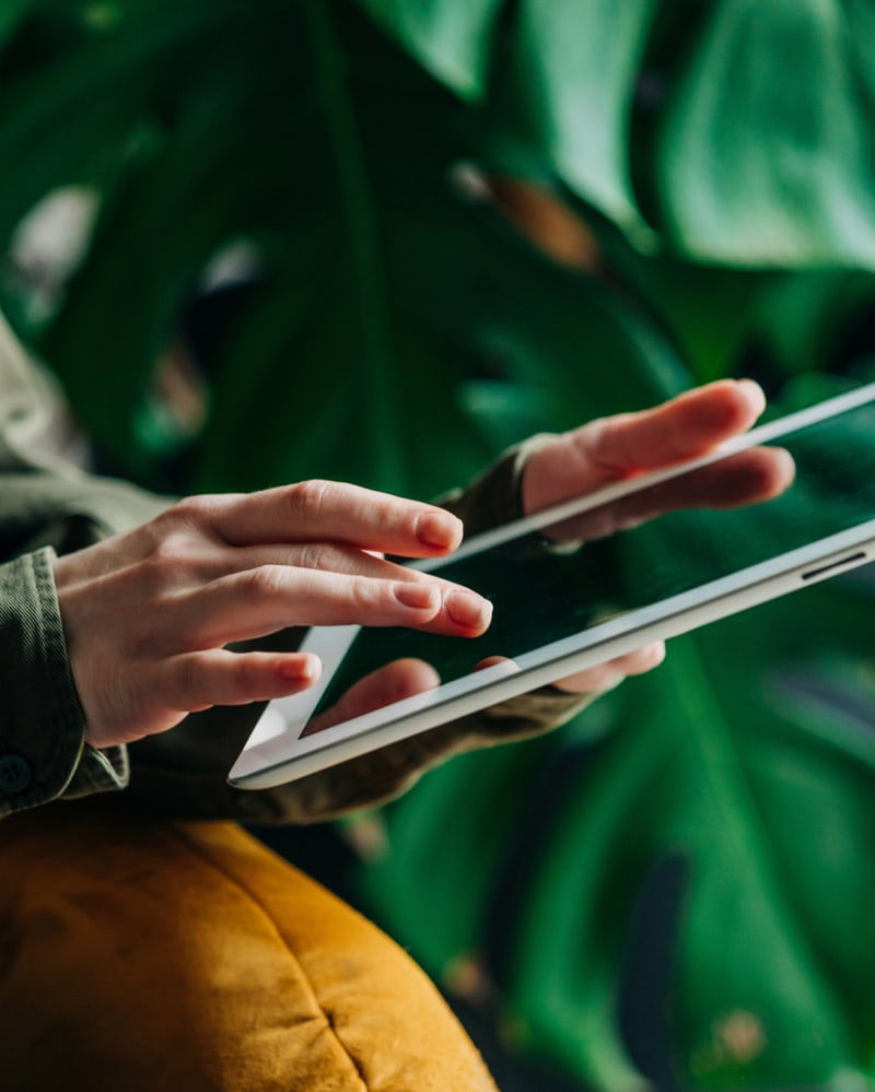 A close-up of a person holding a tablet with a large plant in the background.