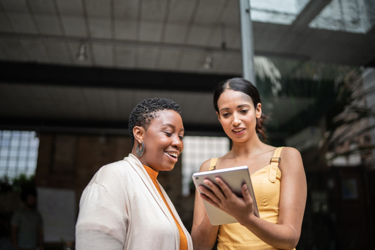 Two smiling women are looking at an image on a tablet in a large conference room. 