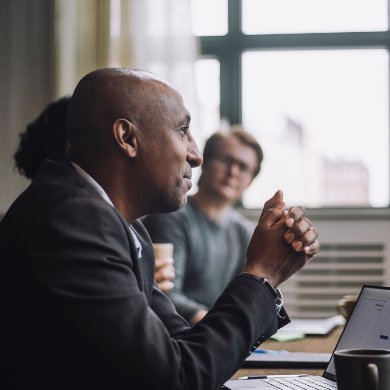 A Black executive wearing a suit attends a meeting with his colleagues in a brightly lit conference room. 