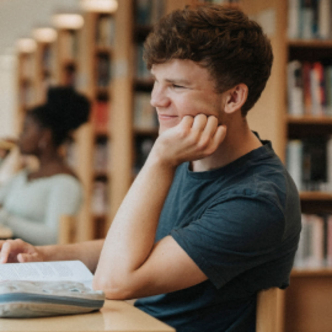 A smiling young man is reading a reference book in a library. 