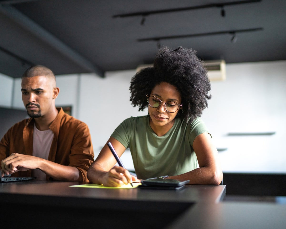 A woman wearing glasses is writing in a notebook, seated next to a man who is typing on a computer.