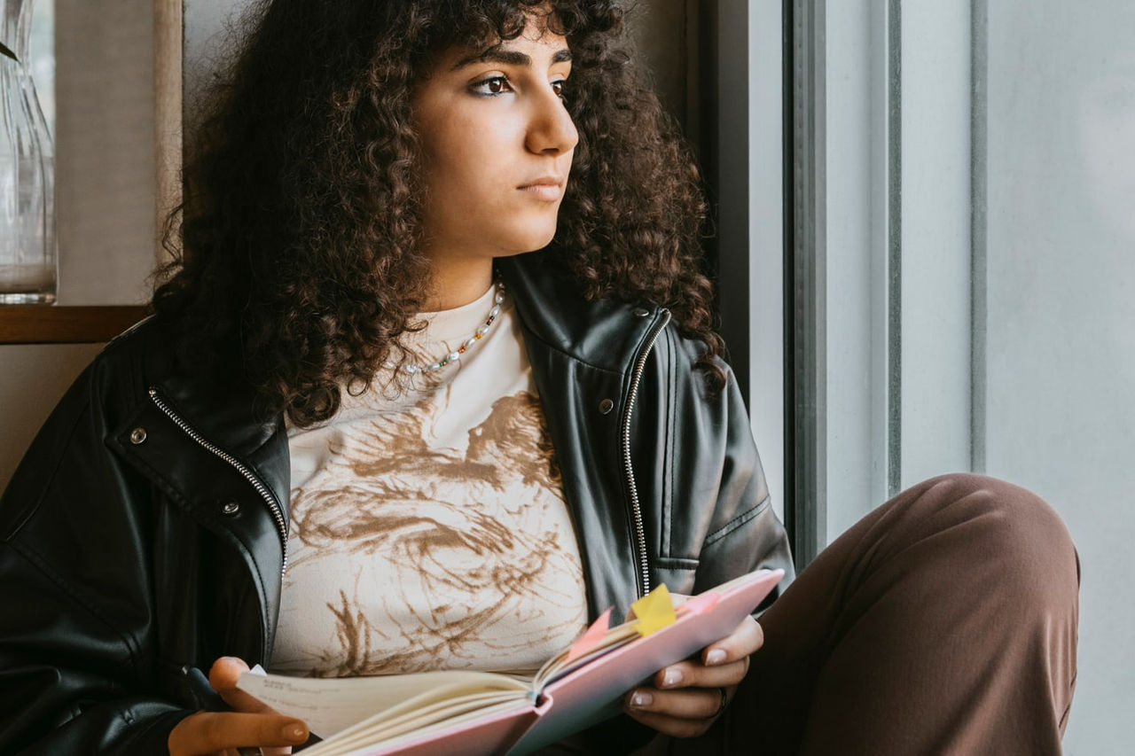 A young woman is seated in a chair with an open book. She has paused in her reading to look thoughtfully out a nearby window.